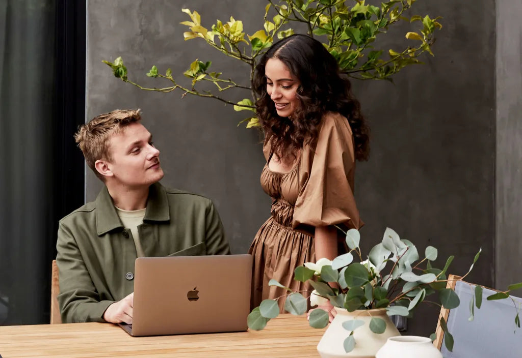 Two people reviewing design ideas together at a table with a laptop, surrounded by indoor plants in a modern studio setting